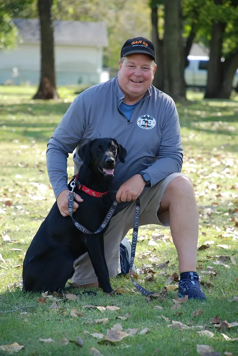 Michael Harvey kneeling on a leaf-covered lawn beside Simon, his black Labrador, both in search-and-rescue K9 gear